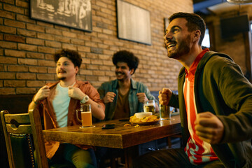 Young excited man and his male friends cheering for their team while watching sports match in pub.