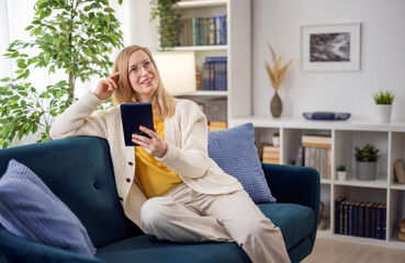 Obraz premium Dreamy caucasian woman in casual clothes and eyewear using digital tablet at home. Mature lady with blond hair sitting on comfy couch and looking aside.