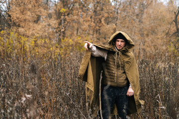 Serious hiker male in raincoat tent standing in thicket of bushes in cold overcast day and pointing finger in direction, looking at camera. Concept of exploration, travel and adventure in nature. - Powered by Adobe