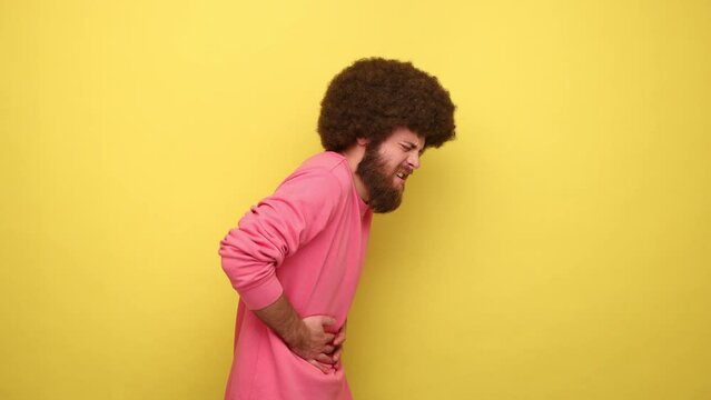 Side view of unhealthy man with Afro hairstyle feeling pain in stomach, suffers diarrhea or constipation risk of appendicitis, wearing pink sweatshirt. Indoor studio shot isolated on yellow background