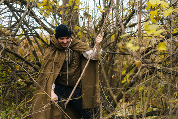 Portrait of smiling tourist male wearing raincoat tent chopping small branches with small shovel in forest on overcast cold day. Concept of scout, research, travel and survival in nature.