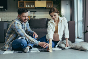 Young couple, man and pregnant woman, playing board game of jang together, sitting on the floor at home, having fun together - obrazy, fototapety, plakaty