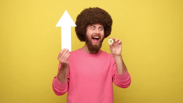 Bearded hipster man with Afro hairstyle showing growth increase of bitcoin, expressing positive emotions, holding white arrow, wears pink sweatshirt. Indoor studio shot isolated on yellow background.