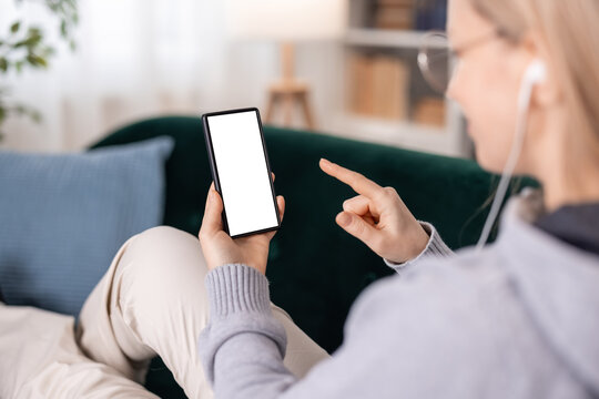 View from shoulder of mature woman in earphones and eyeglasses using smartphone with white mock up screen while resting on sofa. Modern gadgets, people and lifestyles concept.