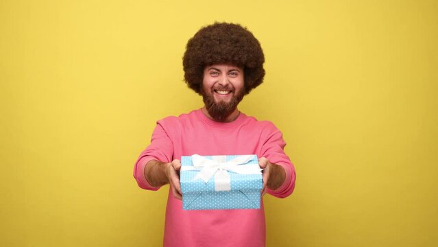 Bearded hipster positive man with Afro hairstyle giving wrapped gift box and smiling at camera, happy holiday, wearing pink sweatshirt. Indoor studio shot isolated on yellow background.