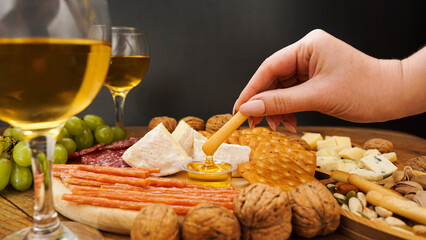 Female hand takes a breadstick and dips it in honey. Cheese and meat platter with assorted cheeses, crackers, nuts on wooden background