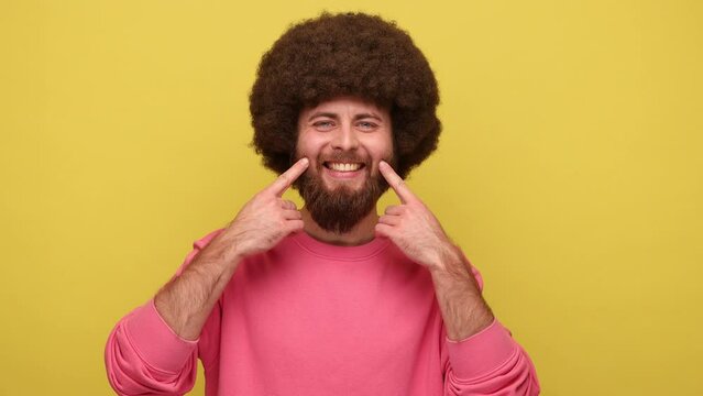 Bearded hipster happy man with Afro hairstyle looking at camera with toothy smile, pointing fingers at his cheeks, wearing pink sweatshirt. Indoor studio shot isolated on yellow background.