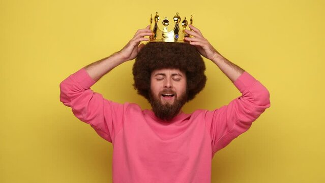Bearded hipster happy self-confident man with Afro hairstyle wearing gold crown, looking at camera with satisfied expression, wearing pink sweatshirt. Indoor studio shot isolated on yellow background.