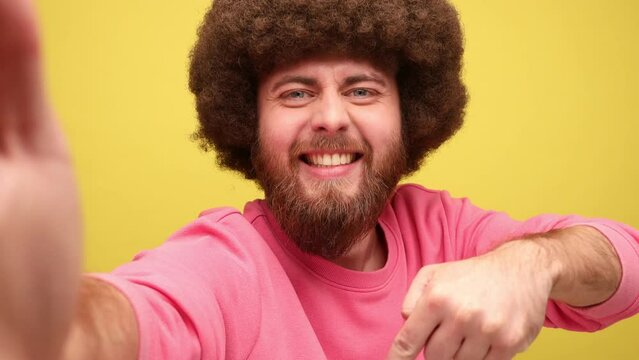 Bearded hipster man blogger with Afro hairstyle broadcasting livestream, pointing down, asking to subscribe, point of view, wearing pink sweatshirt. Indoor studio shot isolated on yellow background.