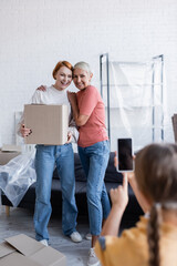 Smiling lesbian couple holding carton box while daughter taking photo on cellphone at home