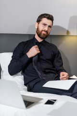 bearded man in eyeglasses adjusting tie and sitting on hotel bed