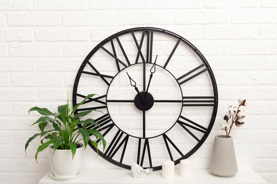 Big large black round clock, green house plant and branch of cotton flowers in vase on table against white brick wall, scandinavian design. Minimalistic composition in pastel neutral tones.