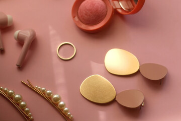 Pink bluetooth earphones, red hairband, pearl hair clips, gold earrings and ring, pink blush and lipstick on pastel pink background. Selective focus.