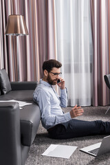 Fototapeta premium bearded freelancer in eyeglasses sitting on carpet and talking on smartphone near laptop and couch
