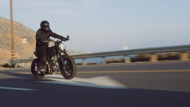 Motorcyclist on beautiful black bike during road trip at seaside town. Picturesque coastal landscape background on a sunny summer day. High quality 4k footage