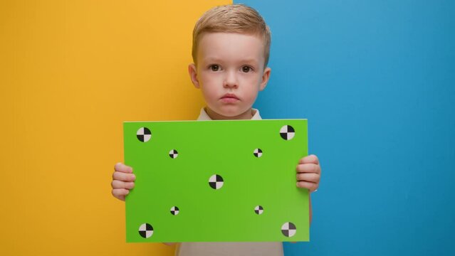 Blonde boy protesting raises banner with green background for keying and with dots for trekking standing on blue-yellow studio background. Protest ukraine, child against, crisis ukraine, ukraine war.