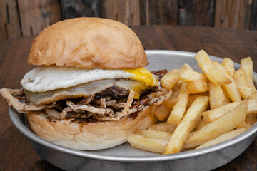 American food rustic presentation. Closeup view of a delicious burger with meat, a grilled egg, crispy onion, provolone cheese, potato bread and french fries, in a metal dish on the wooden table. 
