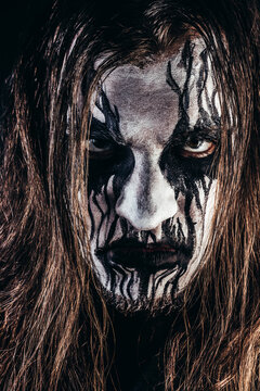 Portrait photo of black metal metalhead man with long brown hair and painted face on black background.