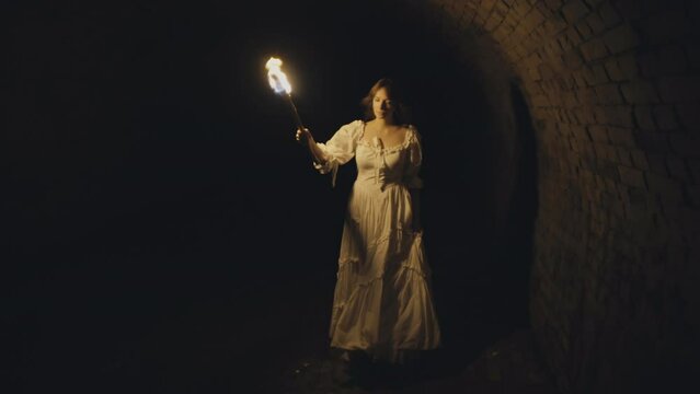 Girl with a torch in a vintage dress walks through a medieval tunnel with brick vaults