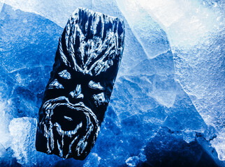 Photo of blue toned ancient wooden fantasy face idol totem laying on cracked ice surface.