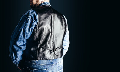 Back view photo of man in jeans shirt and leather biker vest standing on dark blue background.