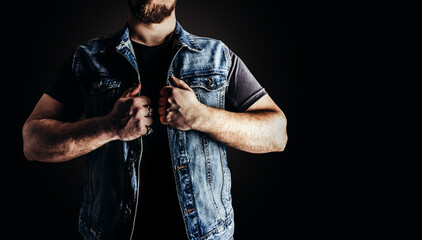 Photo of man in jeans shirt and denim biker vest standing on black background.