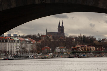 Vysehrad Cathedral under the arch of a stone bridge. The dark cathedral and the whole neighborhood around it is framed by the vault of the stone bridge in the foreground. The river separates the monum