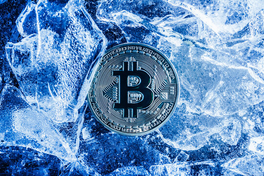 Photo of blue toned bitcoin coin laying on cracked ice surface.