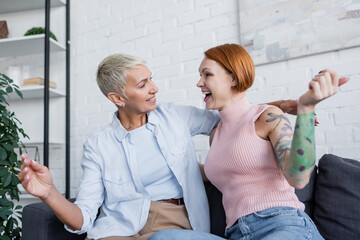 excited lesbian women looking at each other while sitting on couch in living room