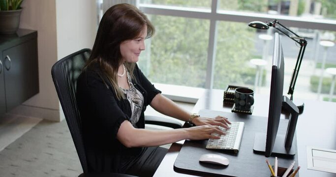 Happy Corporate Brunette Woman Smiling and Typing on a Computer Next to an Office Window - 35mm Medium Tripod Shot 4K