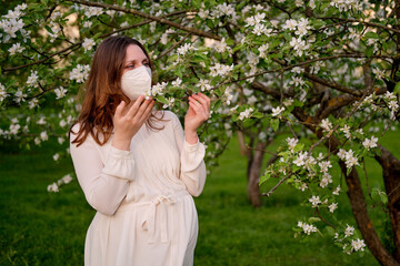 Pregnant woman in medical face mask n95 with apple tree flowers in spring nature. Happiness of pregnancy during covid epidemic, coronavirus pandemic quarantine