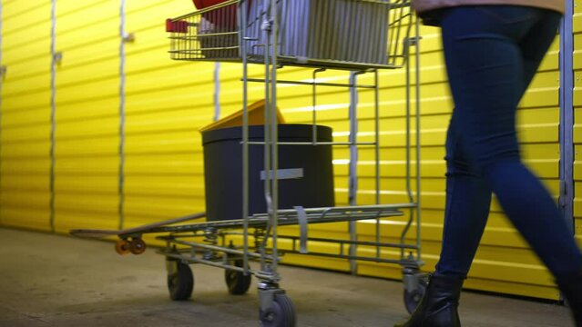 Row of yellow storage rooms doors with legs of young woman passing pushing trolley. Unrecognizable female tenant walking in warehouse self storage indoors leaving
