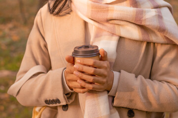 Fashionable young woman holding a takeaway coffee, pumpkin spice latte, in a park on a fall day, hands closeup.