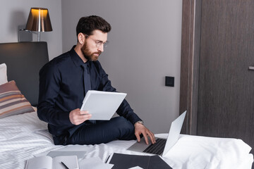 bearded man in suit and eyeglasses using laptop near folders on hotel bed