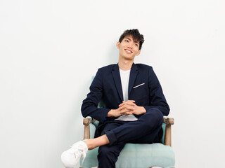 Portrait of handsome Chinese young man in dark blue leisure suit sitting in armchair and posing against white wall background. Hands clasped, leg crossed and looking away, looks confident.