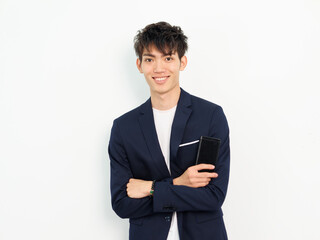 Portrait of handsome Chinese young man in dark blue leisure suit posing against white wall background. Arms crossed with mobile phone in hand and looking at camera, looks confident.