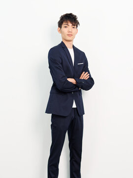 Portrait of handsome Chinese young man in dark blue leisure suit posing against white wall background. Arms crossed and looking at camera, looks confident.