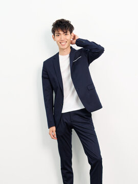 Portrait of handsome Chinese young man in dark blue leisure suit posing against white wall background. Hand on head and smiling at camera, looks confident.