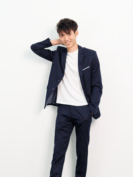 Portrait of handsome Chinese young man in dark blue leisure suit posing against white wall background. Hand in pocket with another hand on head and smiling at camera, looks confident.