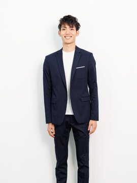 Portrait of handsome Chinese young man in dark blue leisure suit posing against white wall background. Hands on body sides and smiling at camera, looks confident.