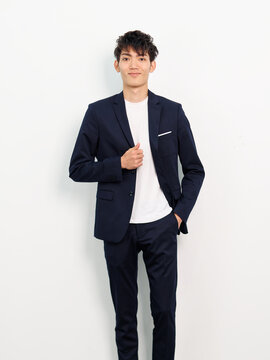 Portrait of handsome Chinese young man in dark blue leisure suit posing against white wall background. Hand in pocket with another hand on suit and smiling at camera, looks smart and confident.