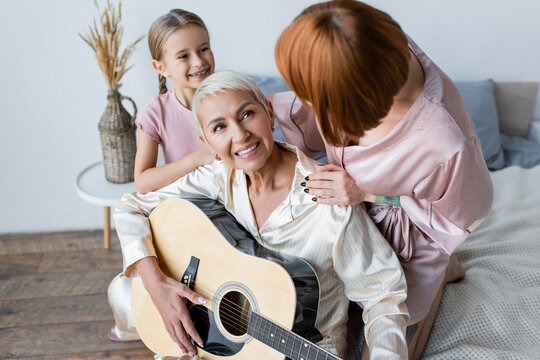 Smiling woman playing acoustic guitar near adopted daughter and girlfriend in bedroom