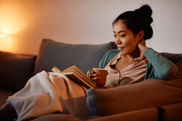 Young Asian woman dinks tea while reading book on the sofa in the evening.