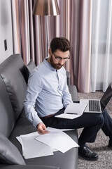 bearded man in eyeglasses holding document and notebook near laptop with blank screen