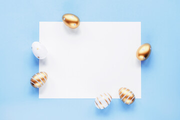 Golden and white painted Easter eggs and blank sheet on a blue background. Top view, flat lay, mock up