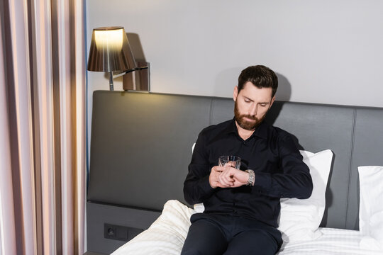 Bearded Man Holding Glass Of Brandy And Checking Time While Sitting In Hotel Bed