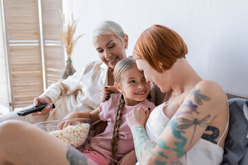 Woman hugging child with popcorn near girlfriend with remote controller at home