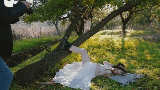 Young cute girl in a white dress posing beautifully in sun glare, lying on the green grass near the tree. the photographer entering the frame takes pictures of the model.