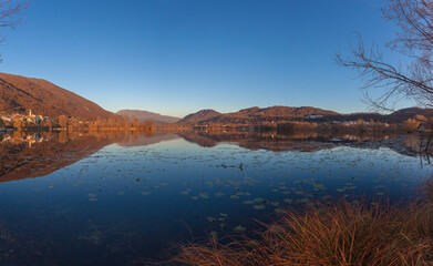 Obraz premium Autumn evening landscape, with a lake nestled between hills and mountains, with a small village of Santa Maria on the shore. Revine Lakes, Veneto, Italy. Concept about reflection and relaxation
