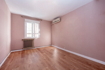 Empty room painted with pastel pink paint tone, wooden floors, white aluminum window and cast iron radiator and air conditioner on the wall